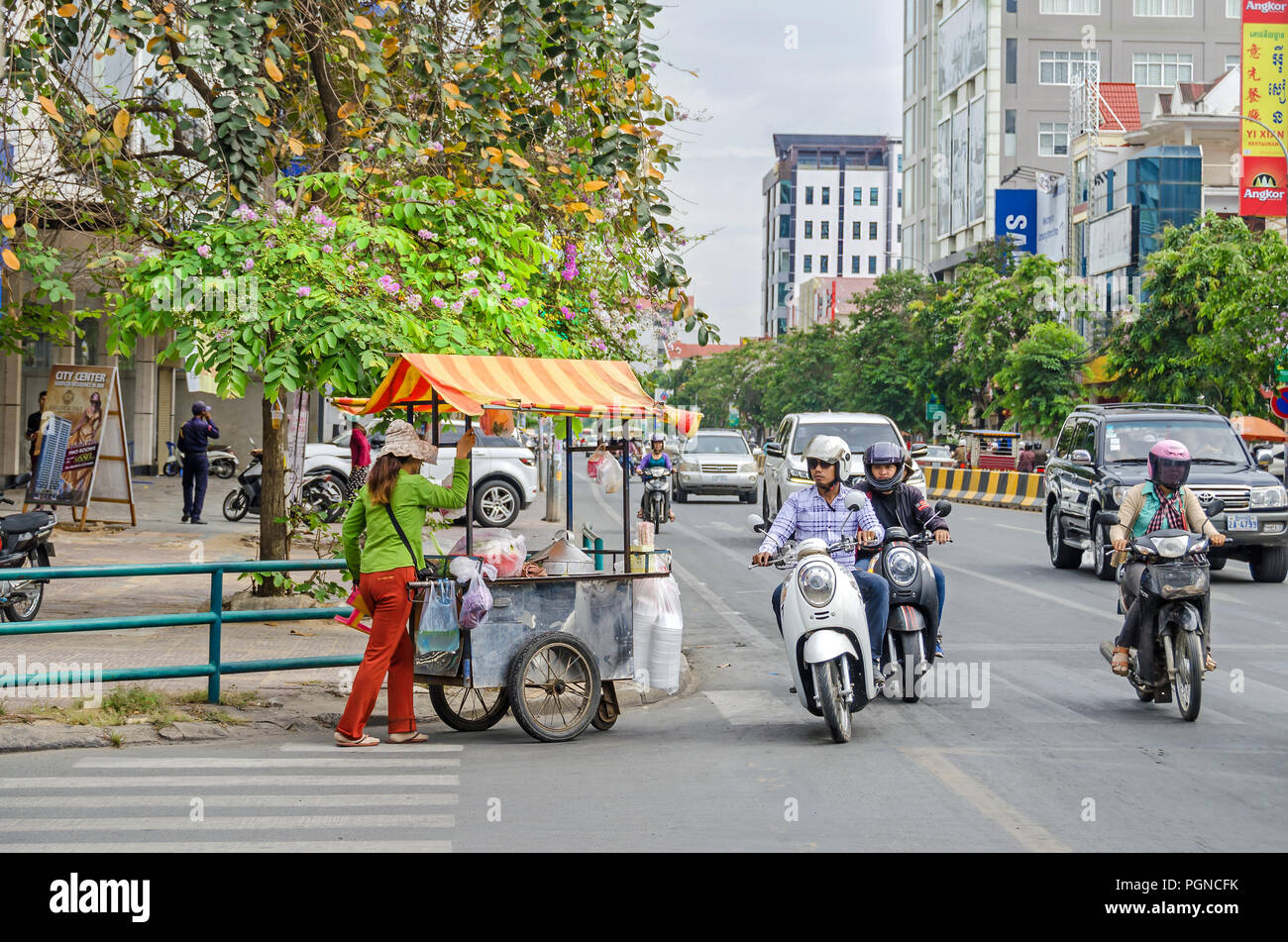Phnom Penh, Kambodscha - April 8, 2018: Blick auf die typisch Schnittpunkt der Kambodschanischen Straßen mit blühenden Bäumen, Mopeds, Autos und einem Straßenhändler Stockfoto