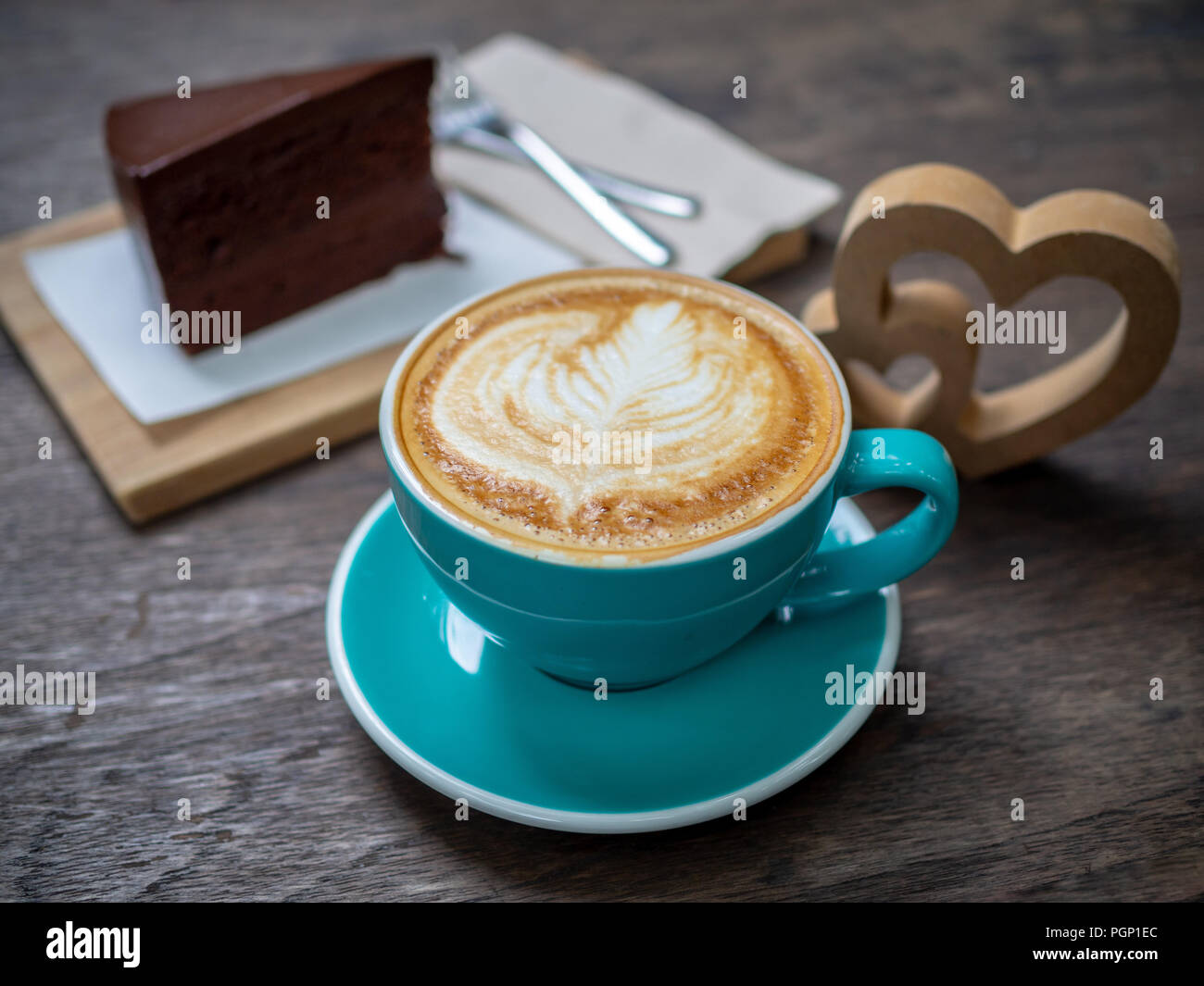 Kaffee latte Art in blau Tasse mit einem Stück Schokolade Kuchen, Gabeln und hölzerne Herz auf hölzernen Tisch Hintergrund. Kaffeeliebhaber Konzept. Stockfoto