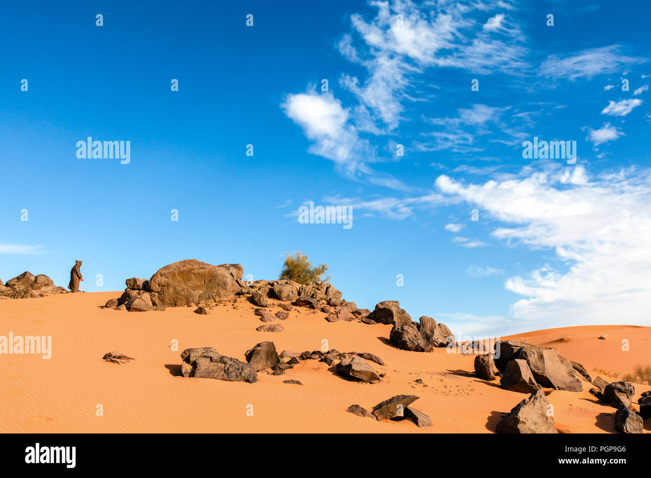 Erg Chebbi, Marokko. Alte Mann mit einem djellaba Robe steht durch einige Felsbrocken in den Sand und in die Ferne schaut. Blue Sky. Platz kopieren Stockfoto