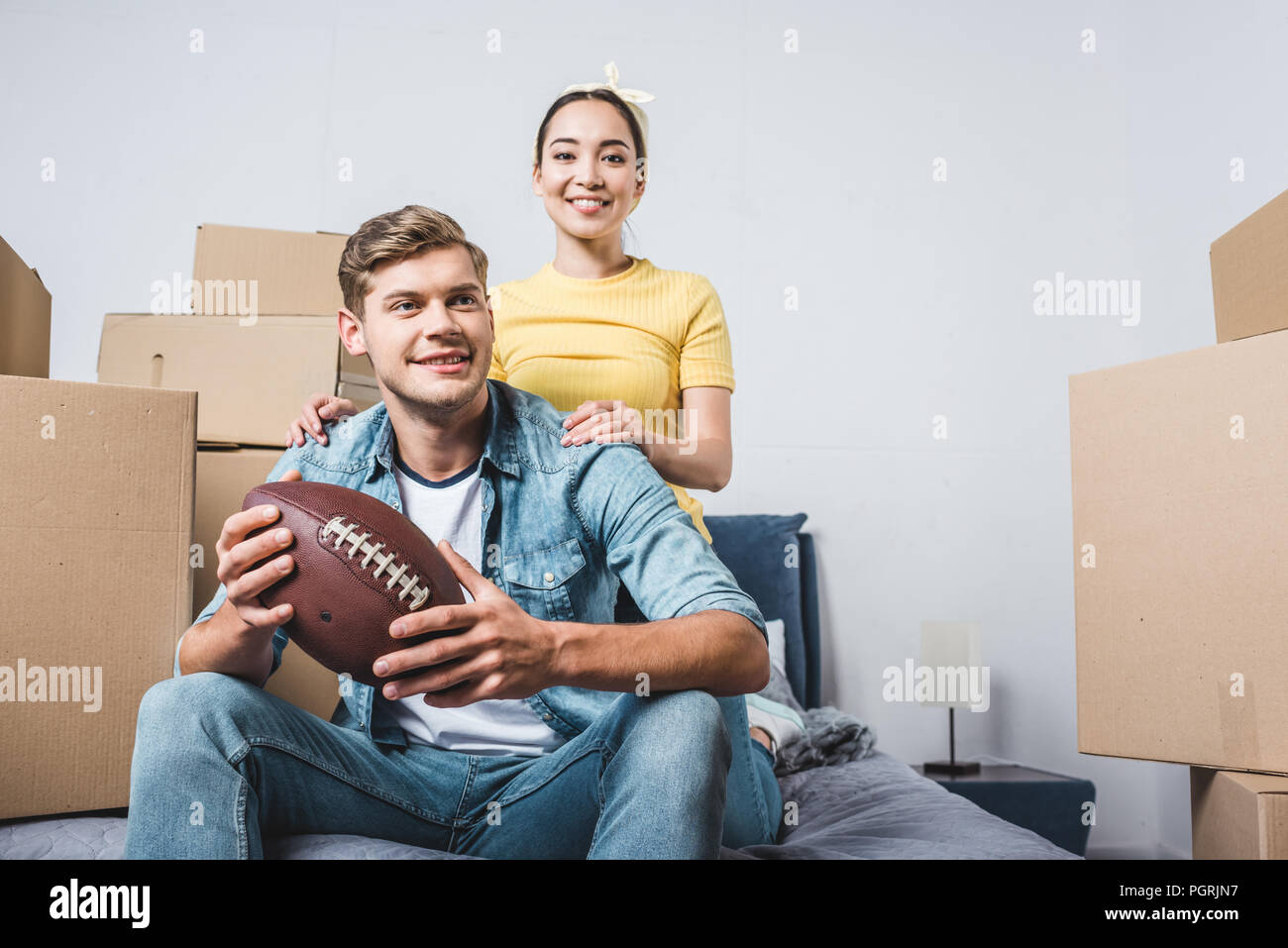 Interracial junges Paar sitzt auf dem Bett mit American football Ball nach dem Umzug in die neue Heimat Stockfoto