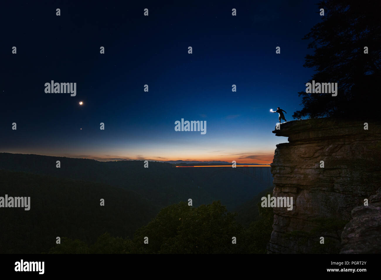 Ein Mann steht gegen den Himmel am Rande einer Klippe mit einem beleuchteten Hand über einem Abgrund gegenüber dem Halbmond in der Nacht. Stockfoto