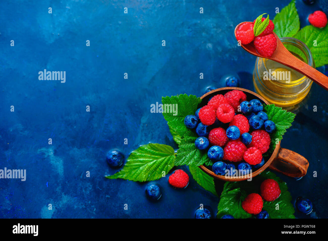 Himbeeren und Blaubeeren in einem keramiktasse mit grünen Blättern und ein Glas Honig. Sommer berry Ernte Konzept auf einem dunkelblauen nass Hintergrund mit kopieren. Ansicht von oben Essen Fotografie Stockfoto