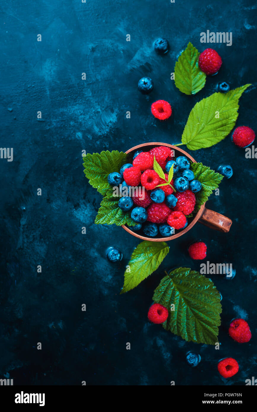 Himbeeren und Blaubeeren mit grünen Blättern in einem keramischen Cup. Sommer berry Ernte Konzept auf einem dunkelblauen nass Hintergrund mit kopieren. Ansicht von oben Essen Fotografie Stockfoto