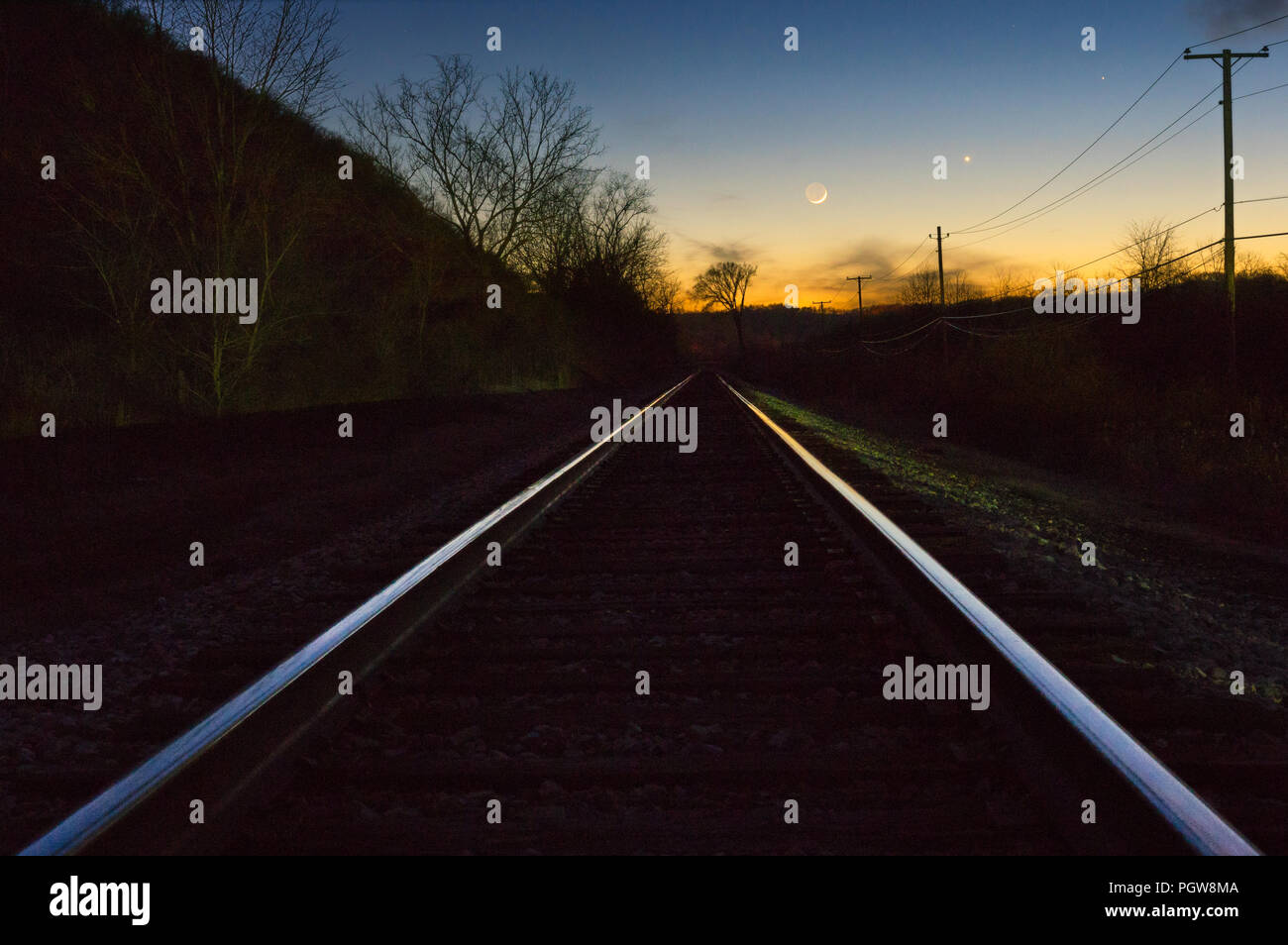 Merkur, Venus, und ein zunehmender Halbmond Linie der Abendhimmel über der untergehenden Sonne und eine Reihe von Eisenbahnschienen. Stockfoto