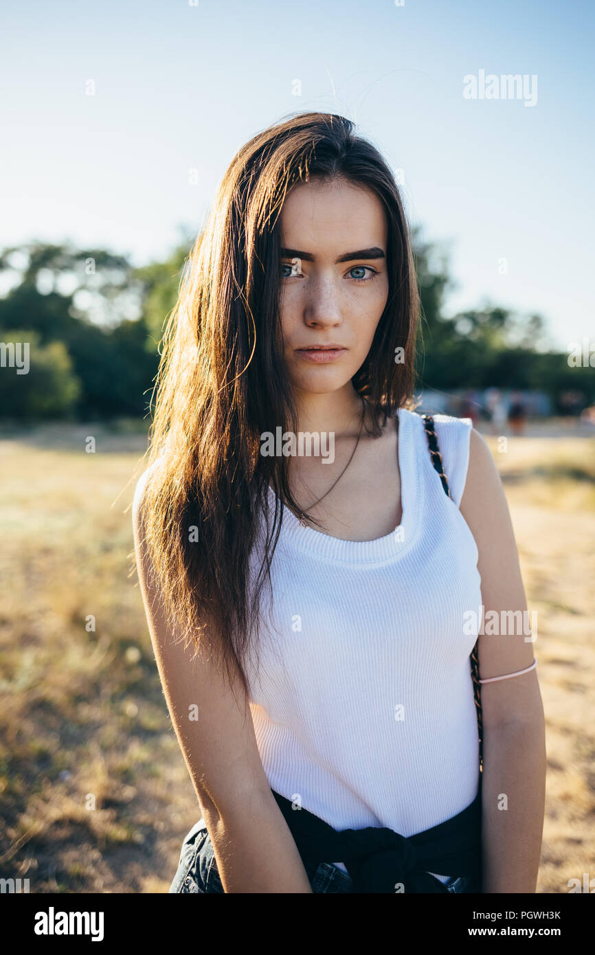 Close-up ehrliches Porträt der Schwere junge Frau mit langen braunen Haaren auf dem Hintergrund der Sommerwiese scheint die Sonne von hinten, vertikale Framing. Stockfoto