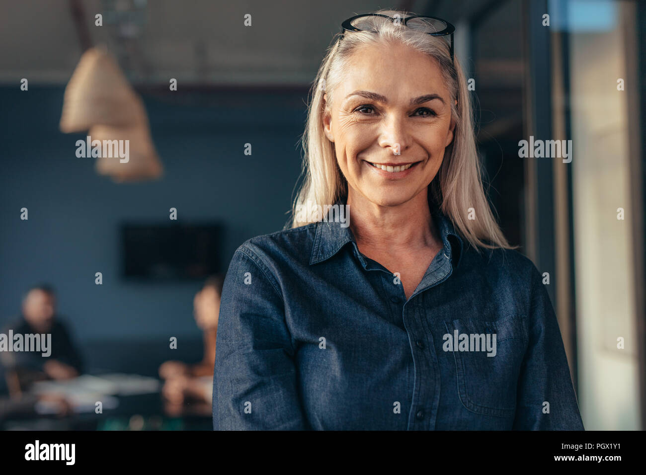 Nahaufnahme von Senior Business Frau im Amt auf Kamera und lächelnd. Reife Frauen im Amt mit dem Team im Hintergrund. Stockfoto