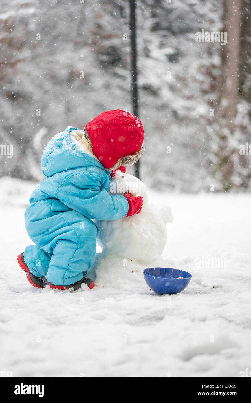 Junge Kind einen Schneemann bauen sich warm eingepackt gegen die Kälte in einem blauen Overall Bücken in der kalten weißen Winter schnee Gestaltung der Kopf, Anzeigen Stockfoto