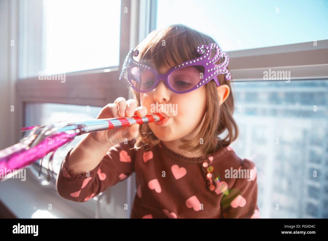 Portrait der kaukasischen Mädchen Kind auf der Fensterbank zu Hause sitzen und Blasen pfeifen Trompete feiert Geburtstag. Kleinkind Vorschulkind tragen Lustig gl Stockfoto