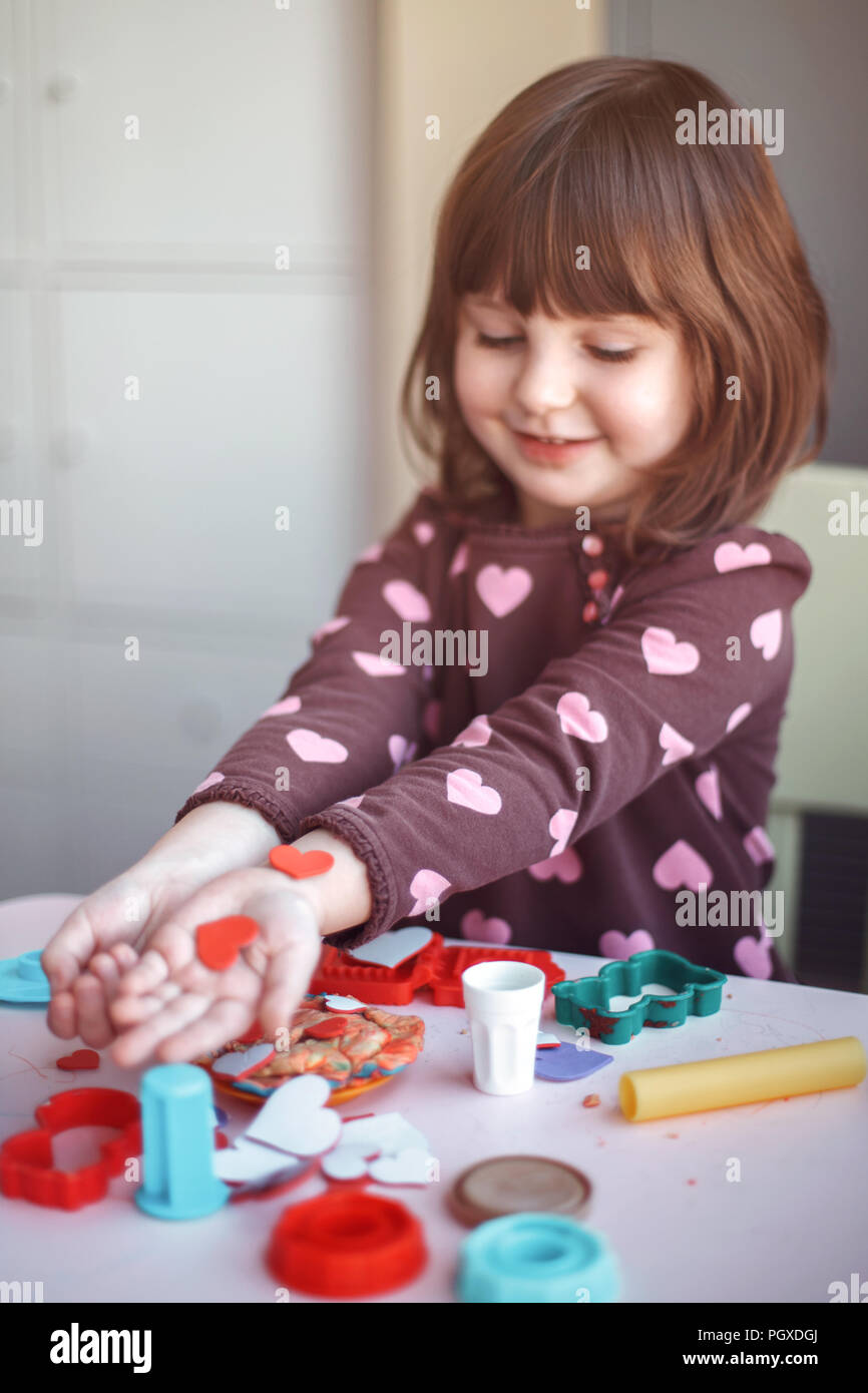 Portrait von niedlichen weißen Kaukasischen Vorschüler Mädchen spielen Plastilin Knetmasse in Innenräumen zu Hause. Kind holding Foam rote Herzen in die Hände. Valentines Tag Stockfoto