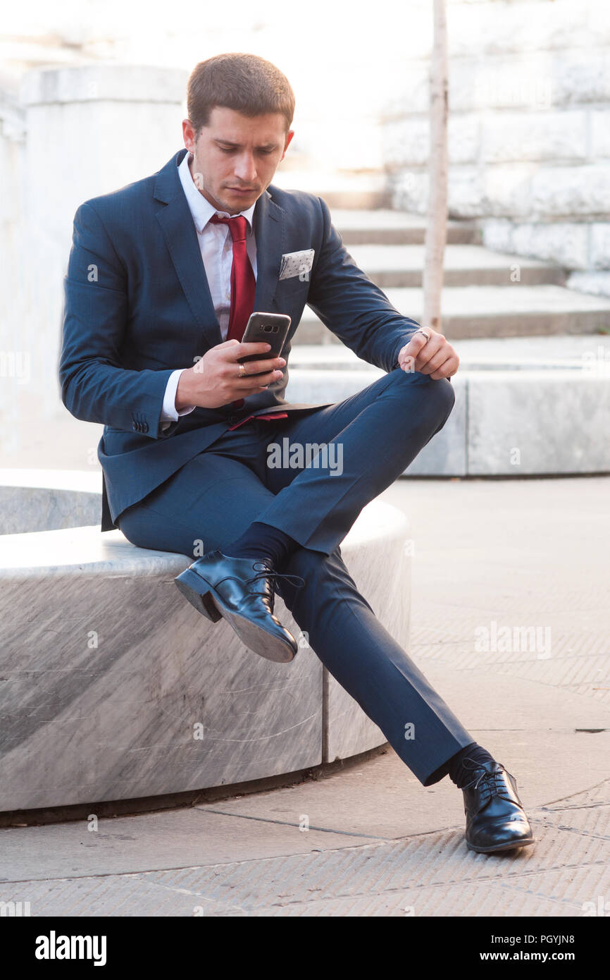Junge gut aussehender Geschäftsmann in Anzug und Krawatte lookink am Smartphone sitzen auf einer Marmorbank im Park Stockfoto