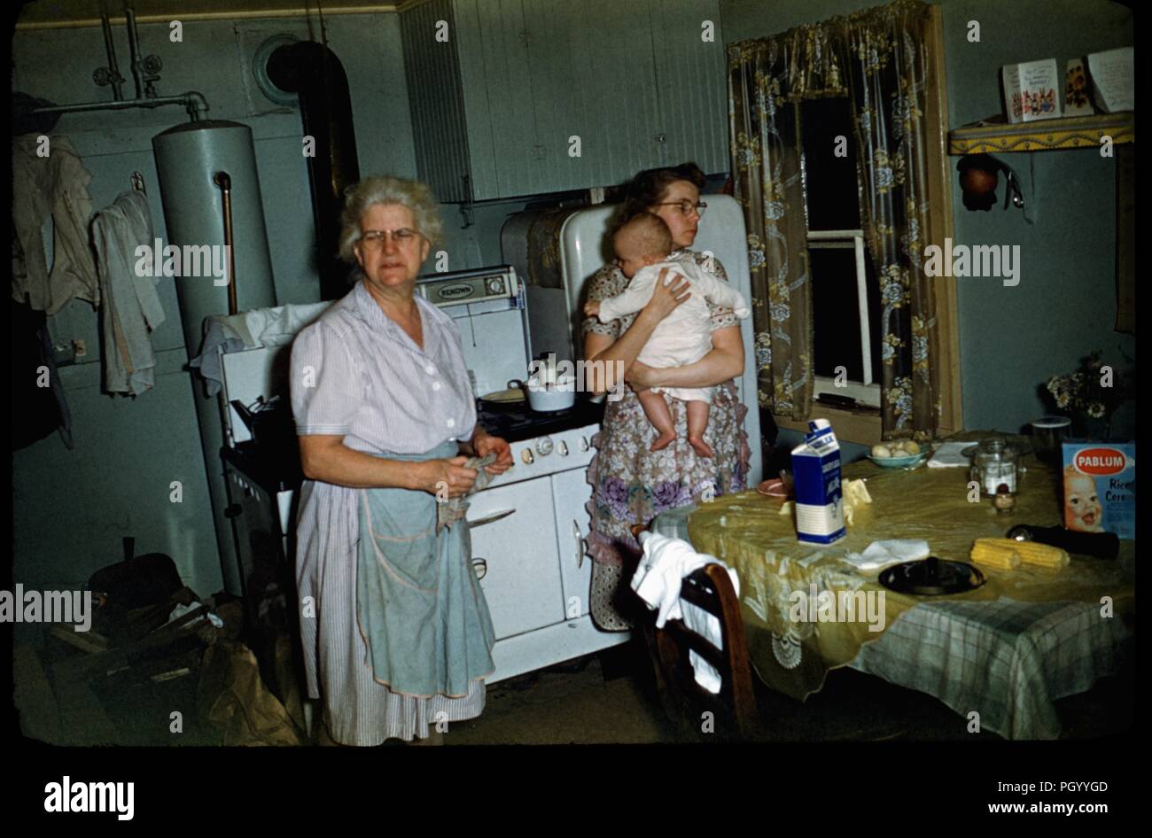 Die junge Mutter hält Ihr Baby und müde erscheinen, stehend in einer Küche in der Nacht mit einer Großmutter in eine Schürze, die Tabelle mit Milch und Pablum baby Müsli, United States, 1955. () Stockfoto