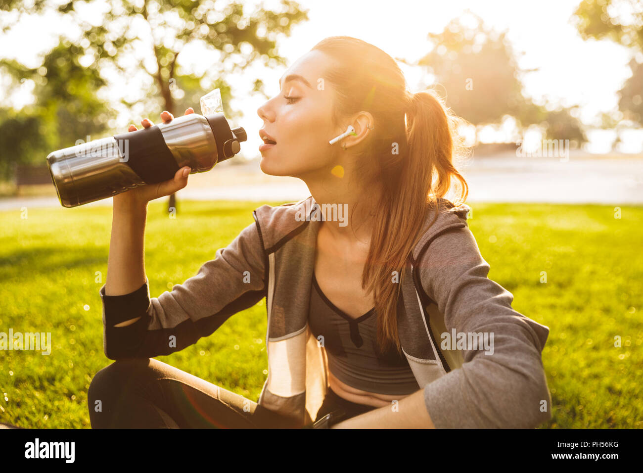 Foto der jungen fitness Frau 20 s in Sportkleidung Trinkwasser aus Metall Schale sitzend auf trainingsmatte im Green Park Stockfoto