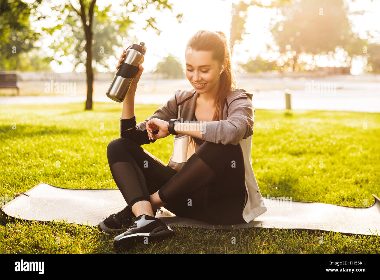Foto von Ihnen fitness Frau 20 s in Sportkleidung Trinkwasser aus Metall Schale und smartwatch sitzend auf trainingsmatte im Green Park Stockfoto