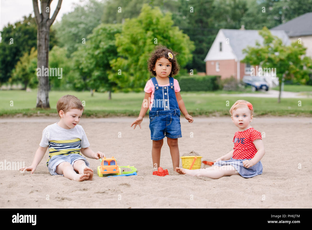 Portrait von drei süße Kaukasischen und hispanische Latein Kleinkinder Babys Kinder sitzen im Sandkasten spielen mit Kunststoff farbenfrohen Spielzeuge. Kleine Mädchen und b Stockfoto
