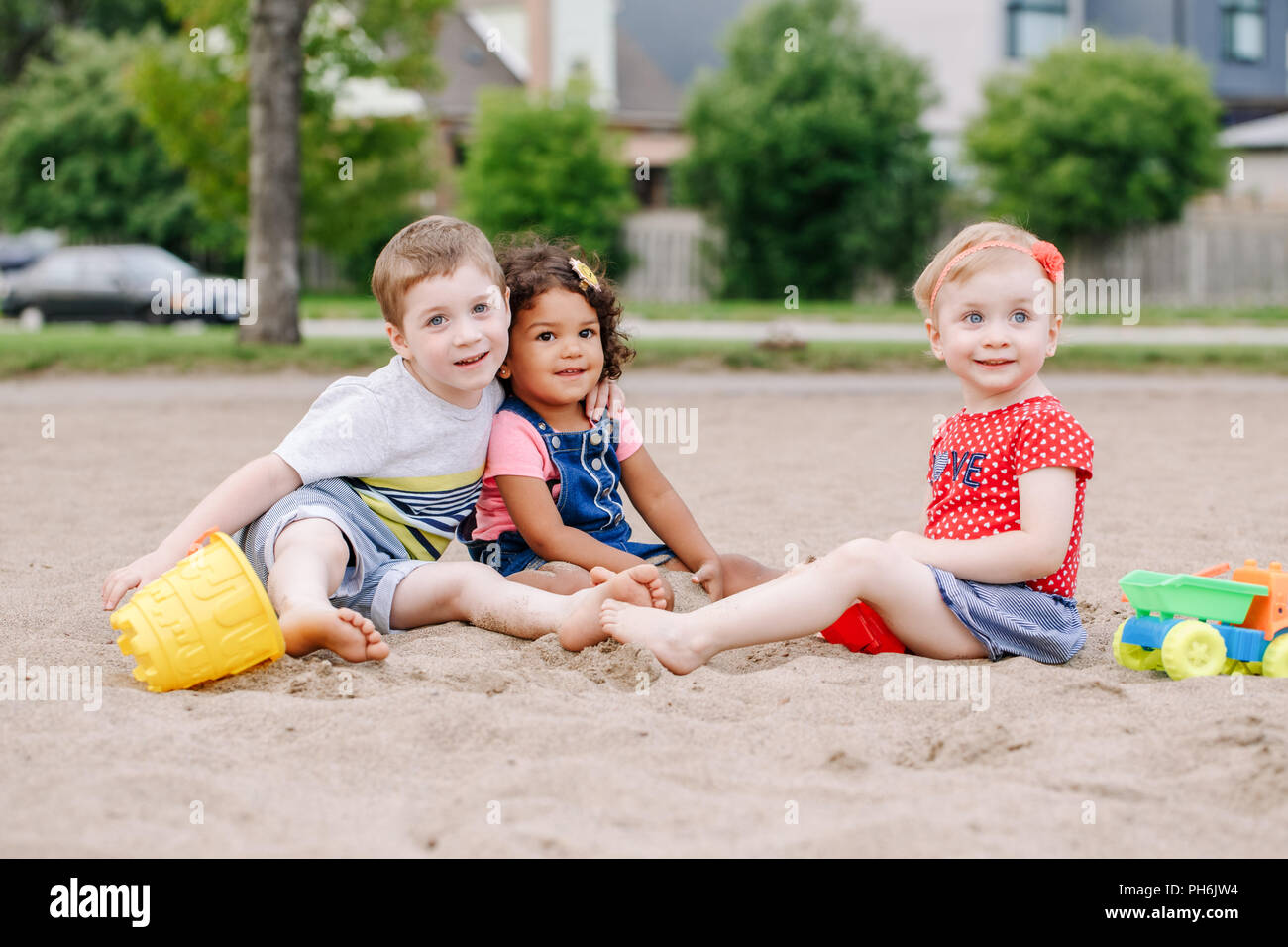 Portrait von drei süße Kaukasischen und hispanische Latein Kleinkinder Babys Kinder sitzen im Sandkasten spielen mit Kunststoff farbenfrohen Spielzeuge. Kleine Mädchen und b Stockfoto