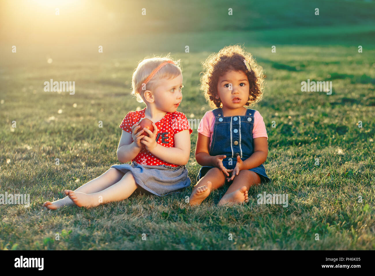 Group Portrait von weissen Kaukasischen und lateinischen Hispanic Mädchen Kinder sitzen gemeinsam gemeinsame Nutzung von Apple. Zwei babys Obst essen draußen im Park im Sommer Stockfoto