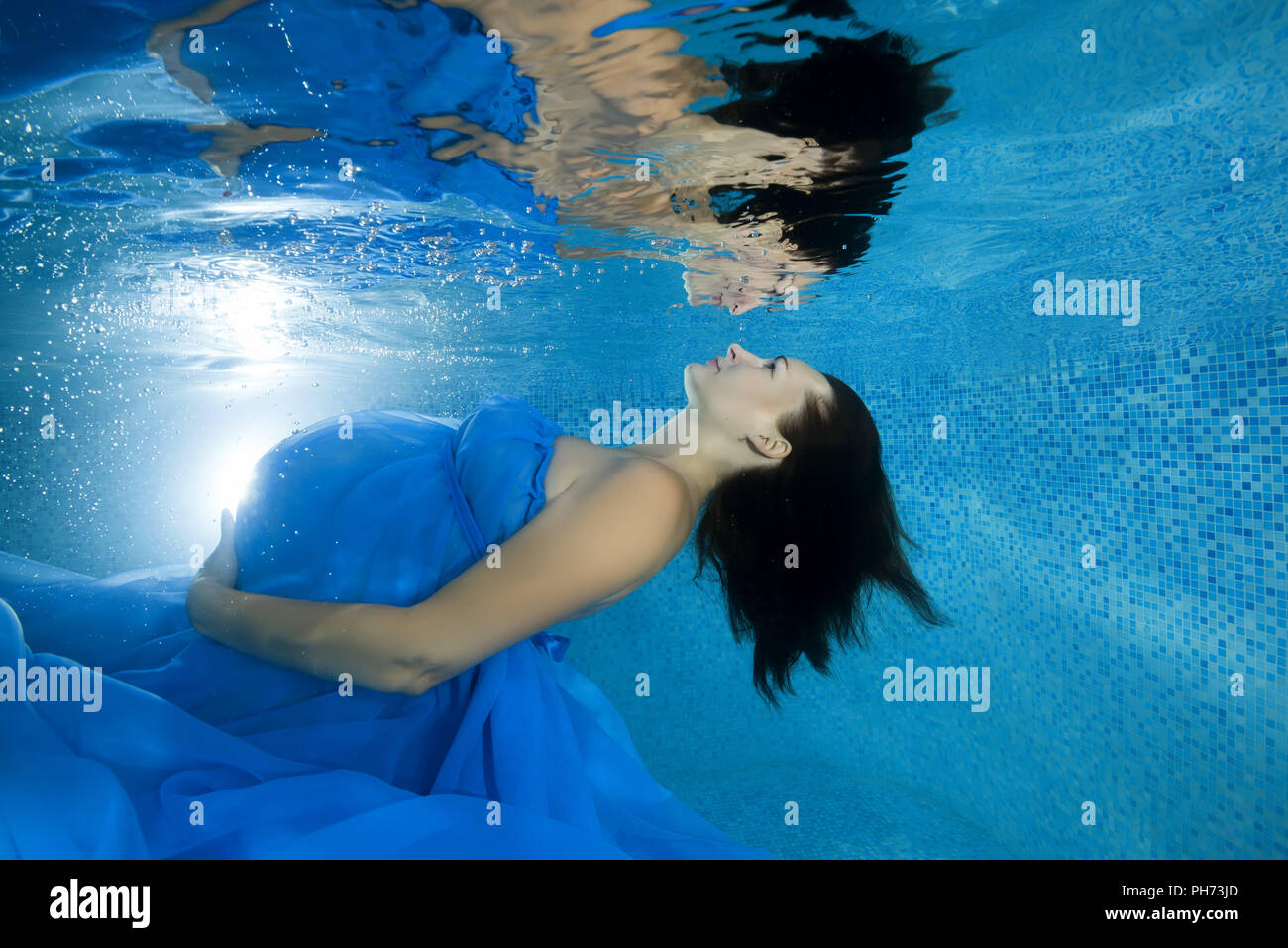 Eine schwangere Frau in einem blauen Kleid schaut sie Reflexion durch die Unterwasserwelt im Pool Stockfoto