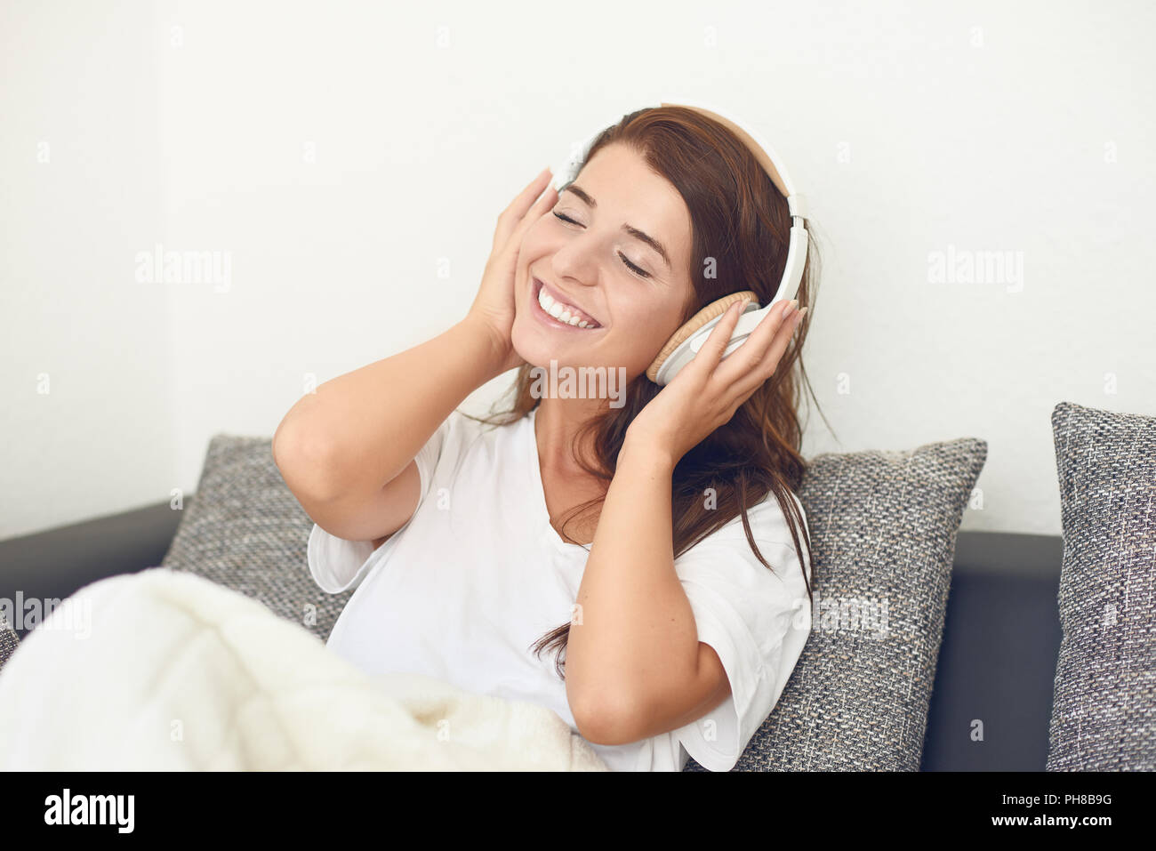 Lächelnd glücklich attraktive quirlige junge Frau auf ein graues Sofa mit einer Decke über die Beine Hören von Musik über Kopfhörer Stockfoto