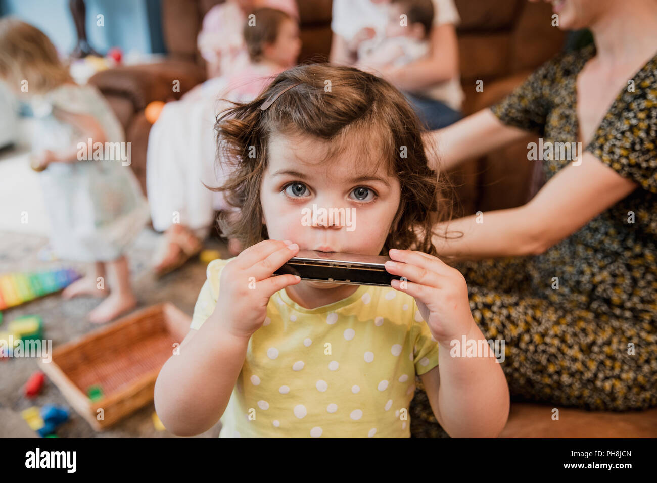 Vorderansicht eines kleinen Kleinkind Mädchen in die Kamera schaut. Sie ist Spaß und das Spielen der Mundharmonika. Im Hintergrund gibt es andere Kinder spielen Stockfoto