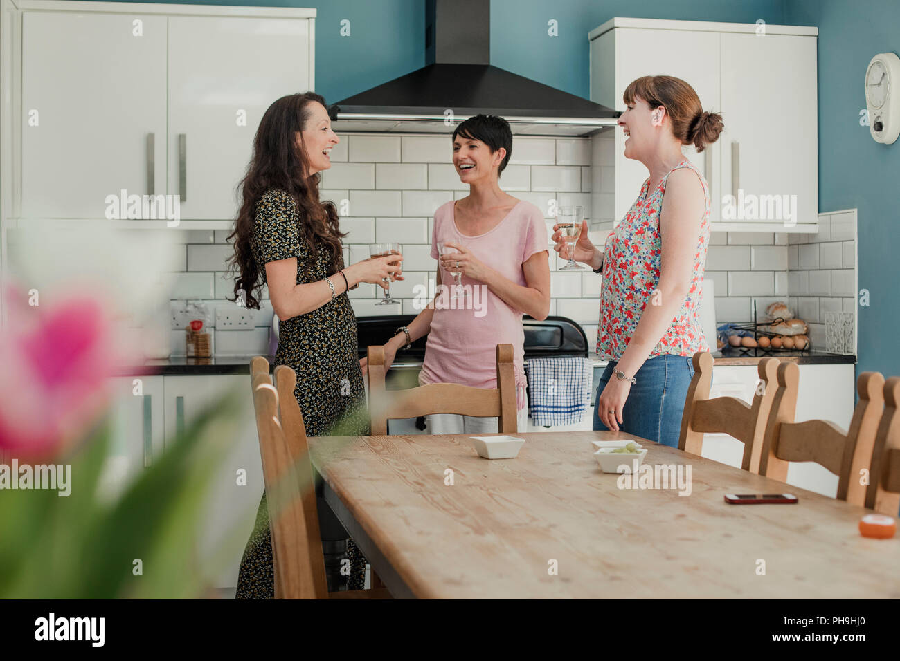 Drei Mitte der erwachsenen Frauen in der Küche bei einem Glas Wein. Die Frauen sind alle ständigen rund um den Herd in der Küche, sprechen und lachen Stockfoto