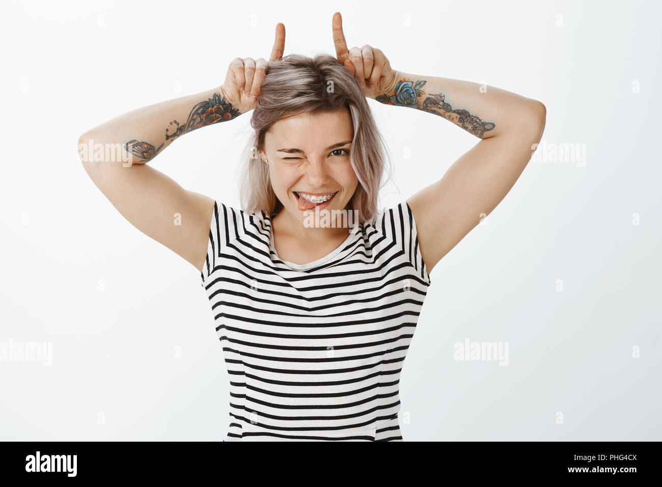 Hartnäckige gut aussehende Freundin mit blondem Haar und Tätowierungen, Zwinkern und breit grinsend, mit niedlichen Klammern auf die Zähne, die Hörner von Zeigefinger auf Kopf, verspielt und flirty Stockfoto