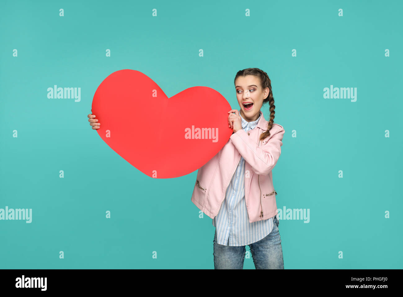 Schöne Mädchen im lässigen Stil, pigtail Haare und Jacke erstaunt stehen und sich zu halten und mit Blick auf Große rote Herzen Form mit überrascht. Indoor, Stockfoto