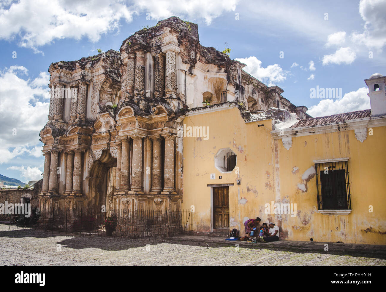 Junge Maya Familie sitzt gegen eine gerissene gelbe Wand der Kirche Ruinen in Antigua, Guatemala Stockfoto