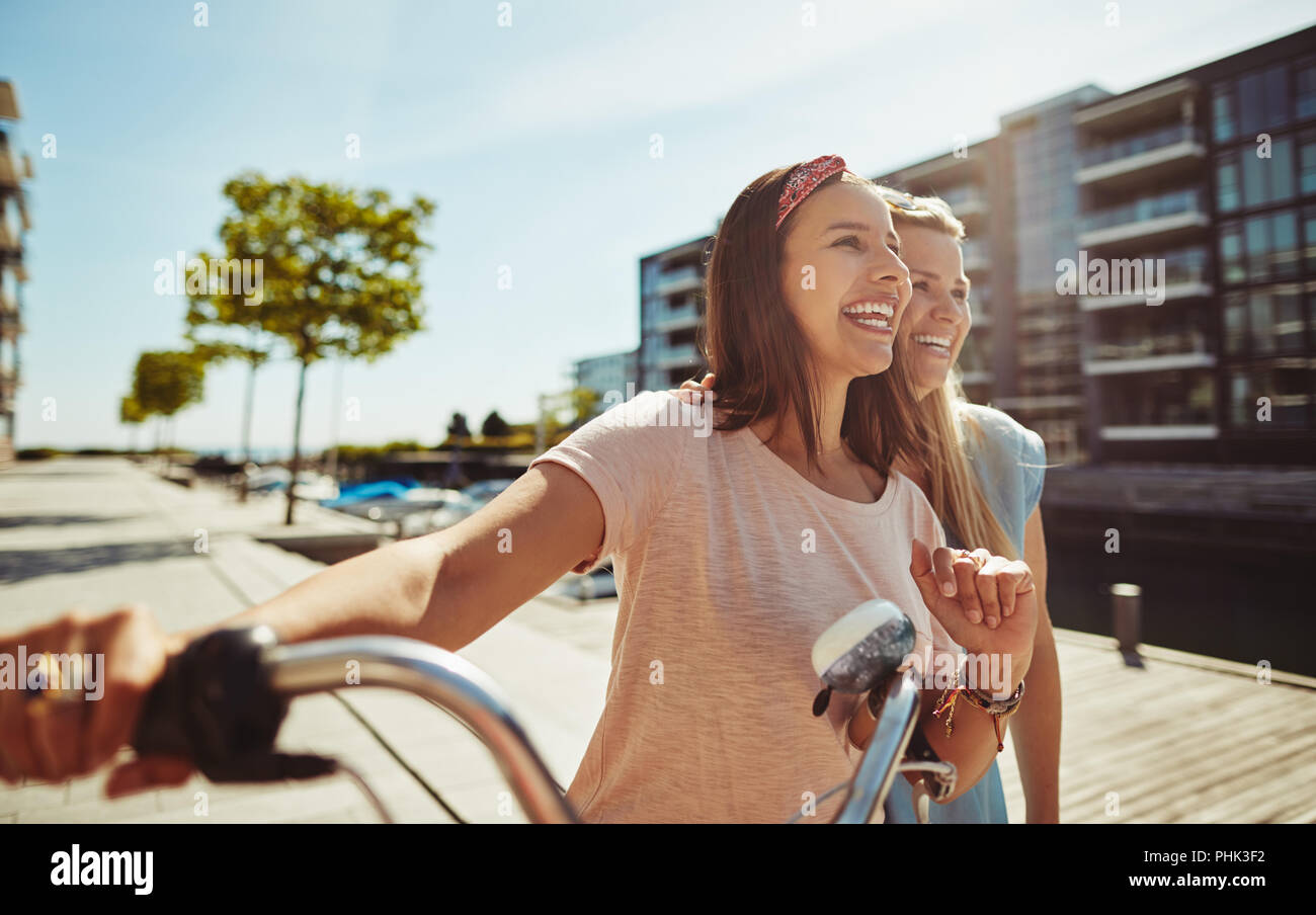 Zwei lachende Junge weibliche Freunde zusammen zu Fuß mit dem Fahrrad durch die Stadt im Sommer Stockfoto