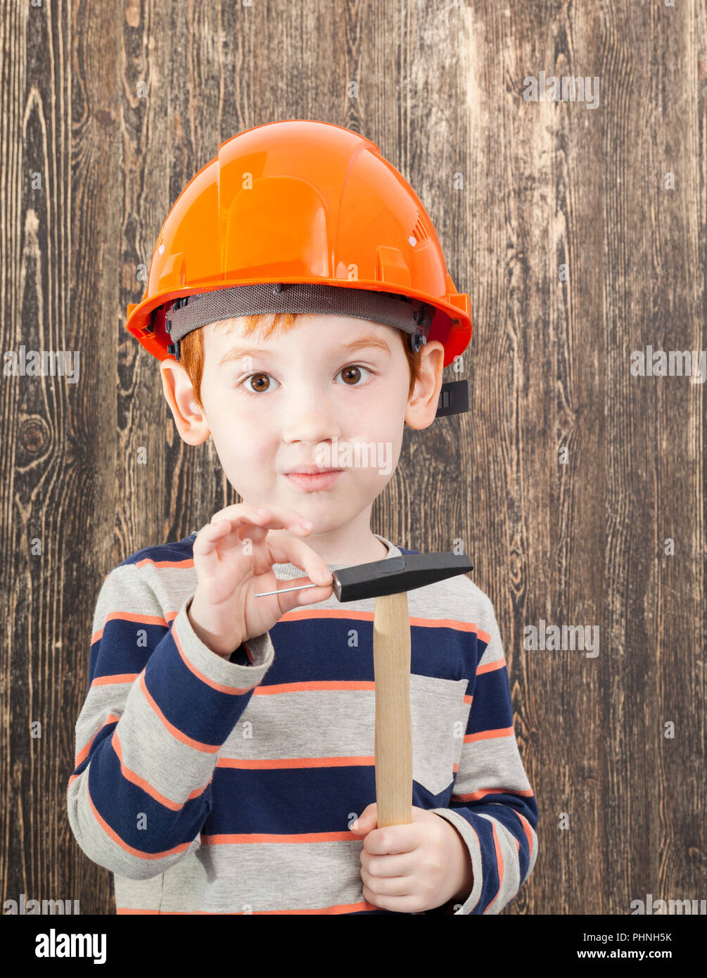 Rothaarige Junge in einem orange Helm mit einem Hammer und einem Nagel, closeup auf schwarzem Holz Hintergrund Stockfoto