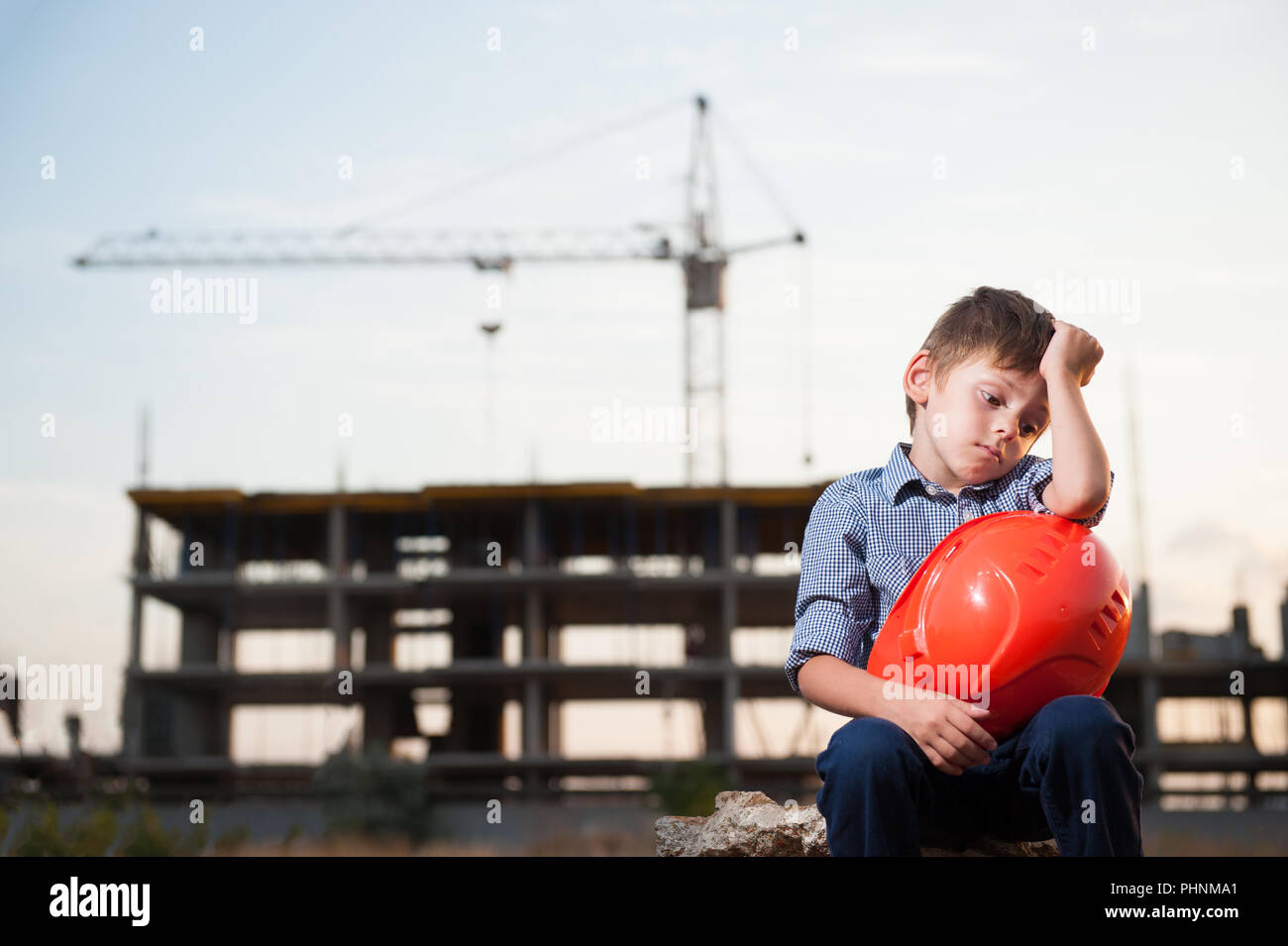 Süß sauer kleiner Junge in der Nähe der Baustelle mit orange Helm in den Händen sitzen Stockfoto