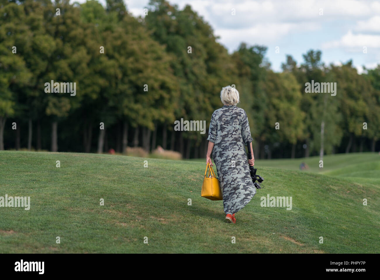 Frau in einem langen Kleid tragen einer Handtasche und die Kamera auf einem Stativ zu Fuß über fein säuberlich geschnittenen Grases auf einem Damm in Richtung Wald Bäume in einer Ansicht von hinten Stockfoto