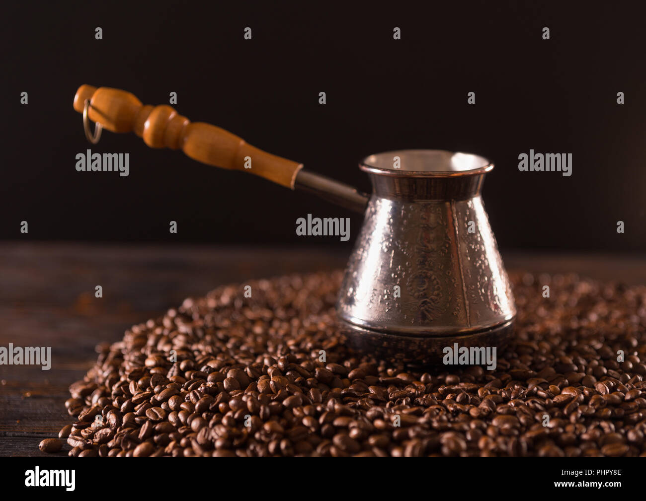 Kleinen Metallbehälter mit langem Griff aus Holz sitzen in Haufen von schwarzen Kaffee Bohnen auf Dunkelbraun Tabelle Stockfoto