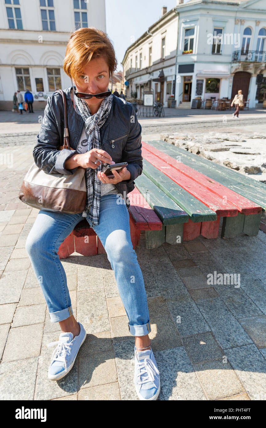 Gegerbt kaukasische Frau mittleren Alters Tourist in Sonnenbrille sitzt und Verwendungen in der Altstadt von Kosice, Slowakei smartphone. Stockfoto