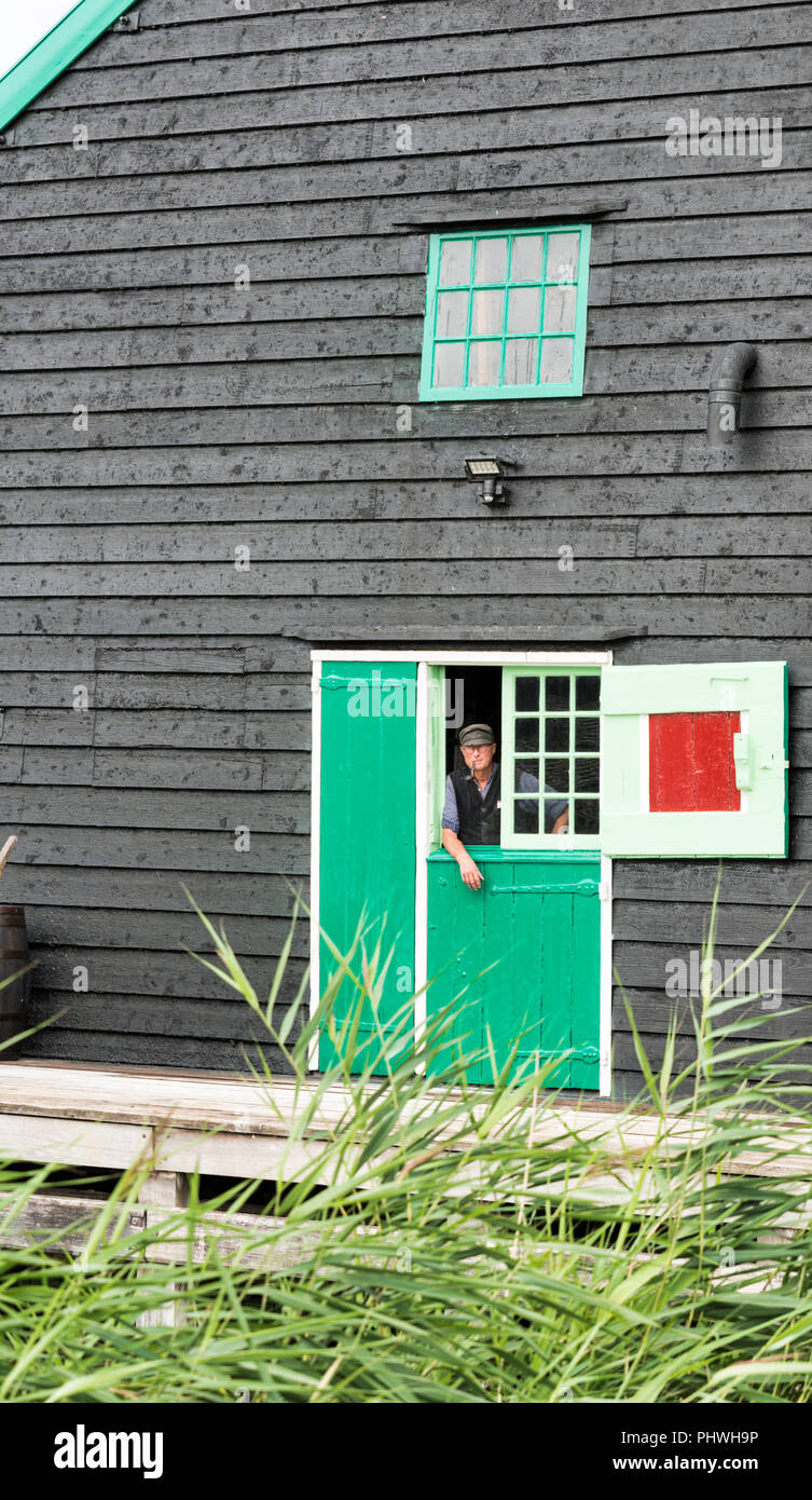 Zaandam, Niederlande, 28-Aug-2018: Niederländische Bauer mit Hut auf und ein Rohr in den Mund schaut aus einem alten Bauernhof mit Fenstern mit Fensterläden, Zaandam ist berühmt für seine alten Bauernhöfen und Leben, wie es war vor hundert Jahren Stockfoto