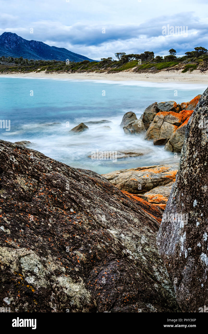 Tagsüber lange Belichtung Erfassung der Wineglass Bay im Freycinet National Park, Tasmanien, Australien, mit orangefarbenen Flechten auf Felsen im Vordergrund. Stockfoto