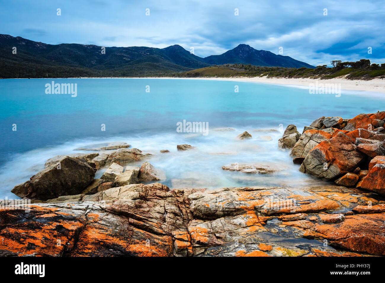 Tagsüber lange Belichtung Erfassung der Wineglass Bay im Freycinet National Park, Tasmanien, Australien, mit orangefarbenen Flechten auf Felsen im Vordergrund. Stockfoto
