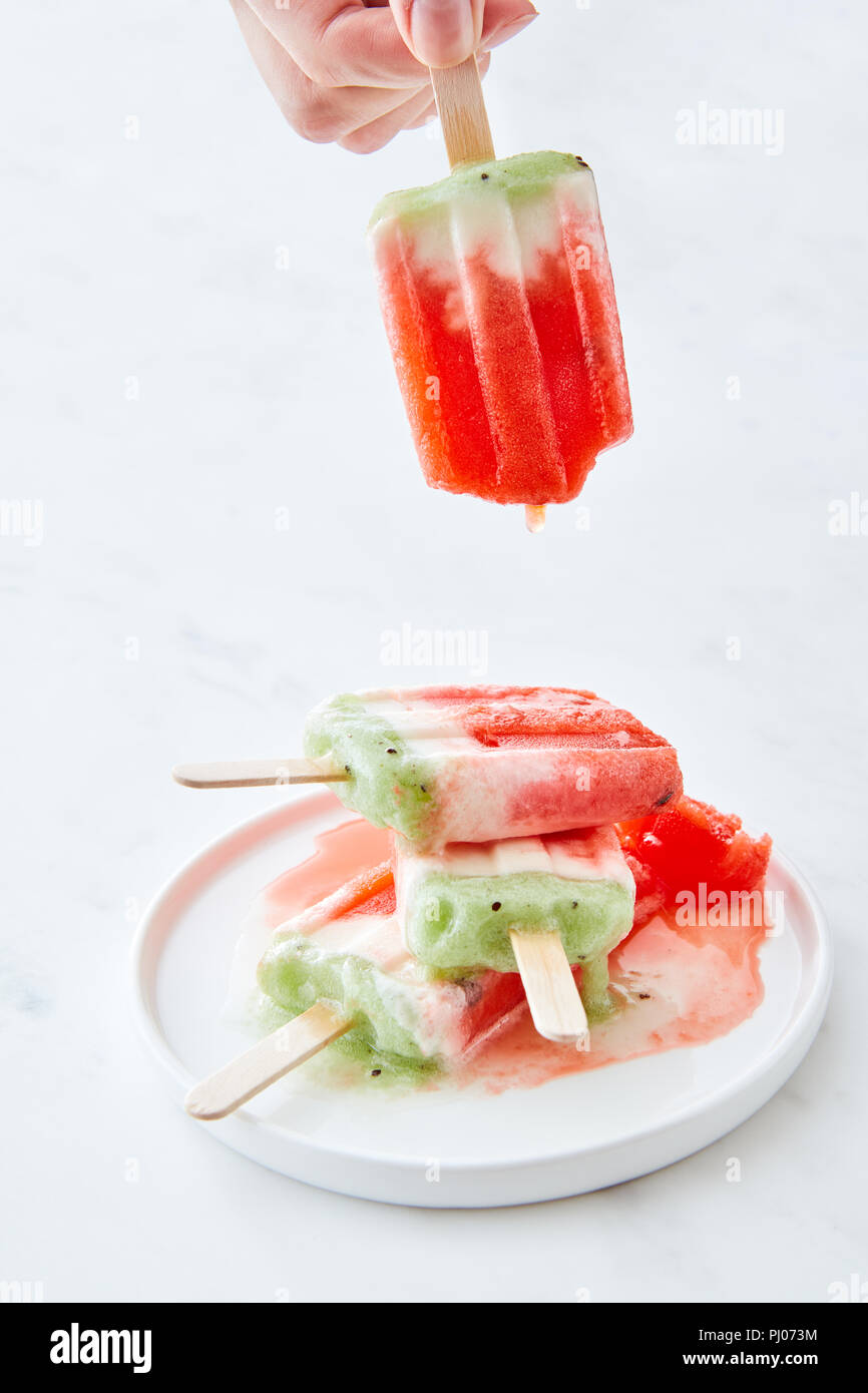 Eis am Stiel in einem Teller mit Spritzern. Hand hält eine Frau eines schmelzenden Eis auf einem weißen Hintergrund mit Kopie Raum Stockfoto