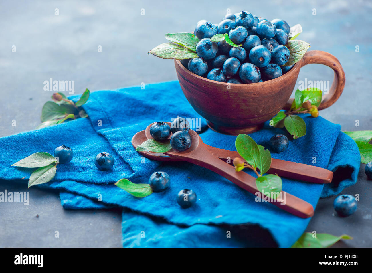 Heidelbeeren im keramiktasse mit holzlöffel und ein Bettwäsche Serviette. Reif und süß Sommer Beeren auf einem Stein. Neutrale Farben, Blau und Grau Stockfoto