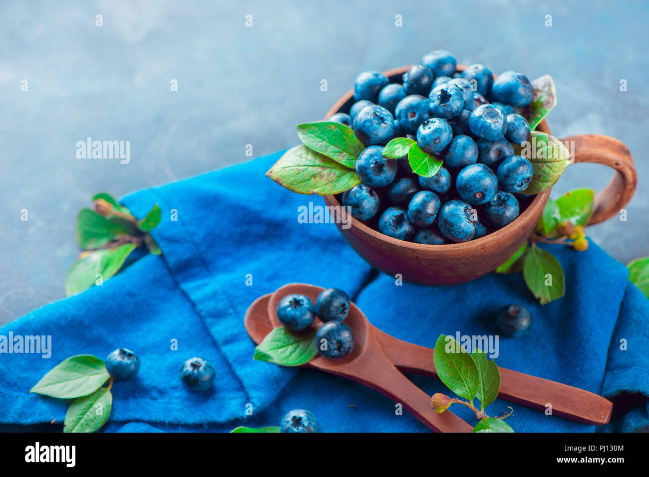 Heidelbeeren im keramiktasse mit holzlöffel und ein Bettwäsche Serviette. Reif und süß Sommer Beeren auf einem Stein. Neutrale Farben, Blau und Grau Stockfoto