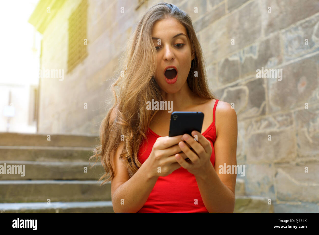 Euphorische junge Frau smart phone aufpaßt, während zu Fuß auf der Straße mit Flair. Überrascht Mädchen mit Smart Phone im Freien. Stockfoto