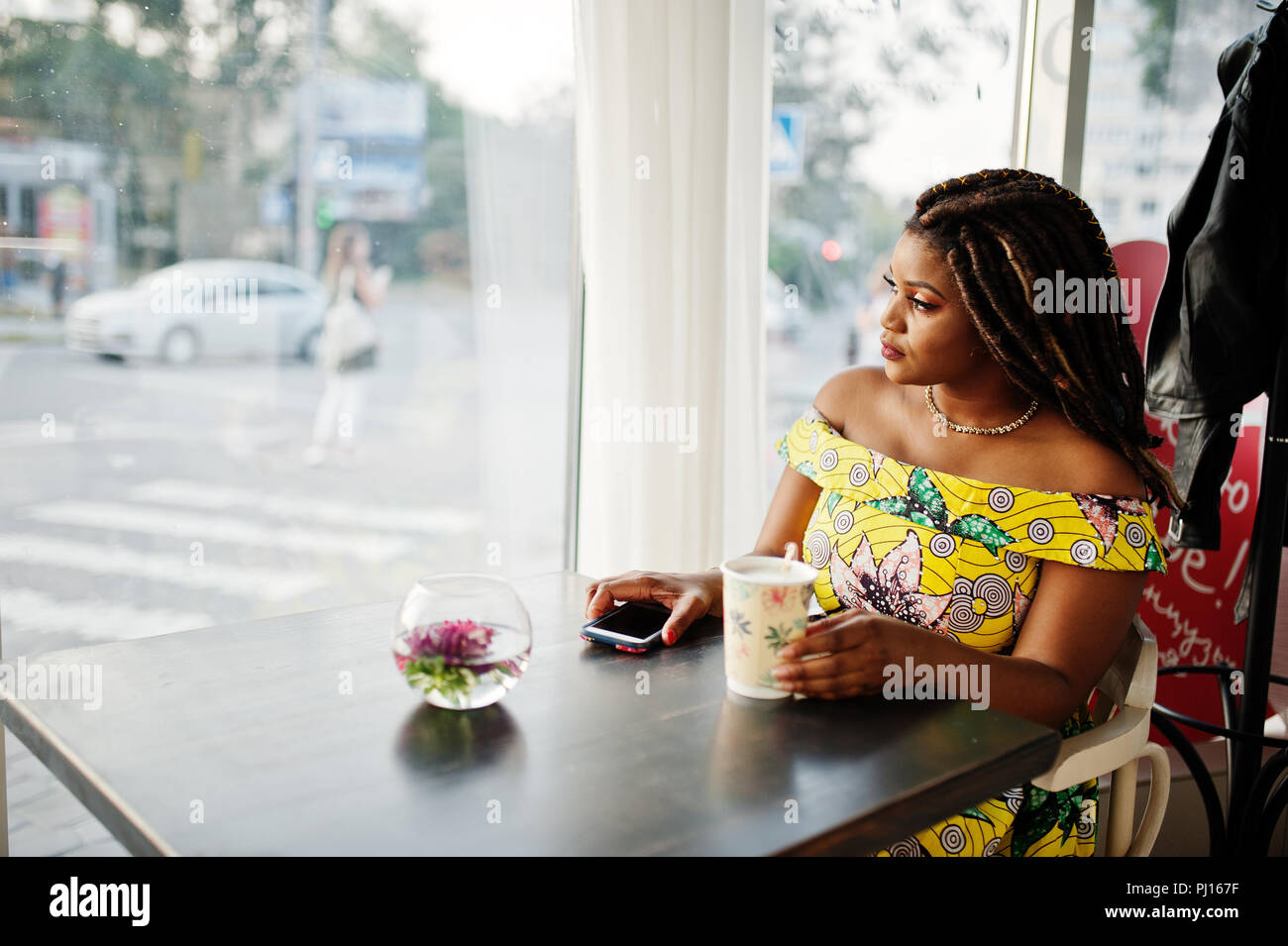 Süße kleine Höhe afrikanische amerikanische Mädchen mit Dreadlocks, Verschleiß an den Farben gelb kleid, sitzen im Cafe bei einer Tasse Kaffee und Fenster. Stockfoto