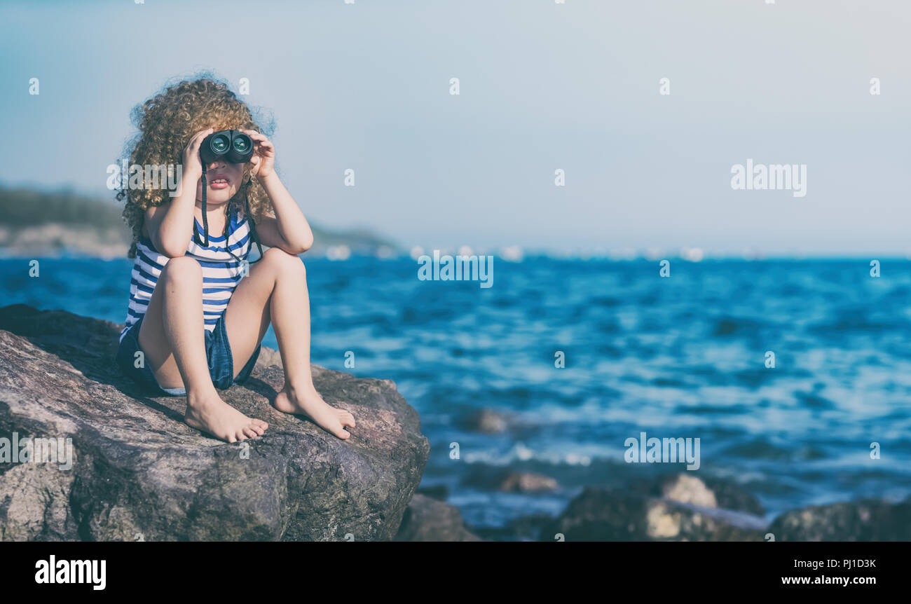 Kleines Mädchen auf der Suche weit weg mit dem Fernglas, sitzt auf einem Felsen in der Nähe eines Meeres Stockfoto