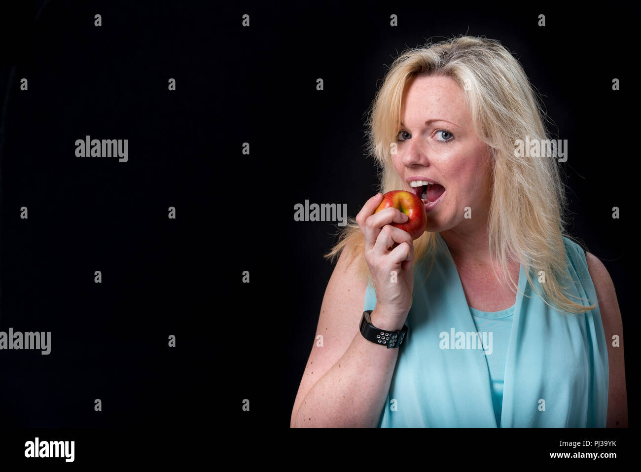 Ausdrucksstarke blonde Frau beißt ein Apple vor einem schwarzen Hintergrund Stockfoto