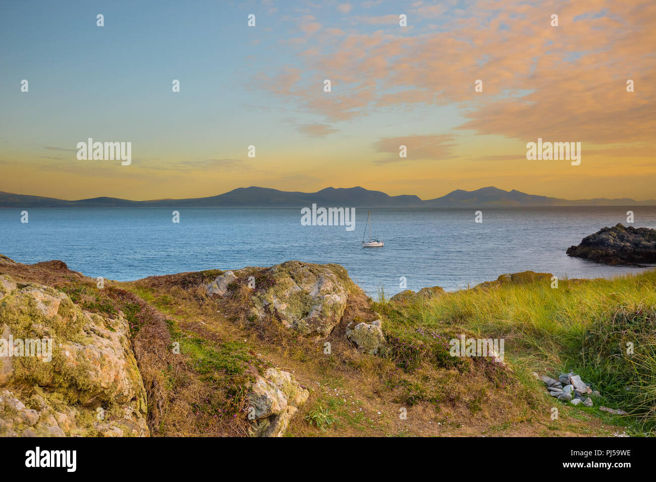 Sonnenuntergangsseelandschaft von Ynys Llanddwyn Headland (Gezeiteninsel in Anglesey, Wales) über Meer in Richtung Yr Eifl (die Rivalen auf Englisch) & entferntes Boot. Stockfoto
