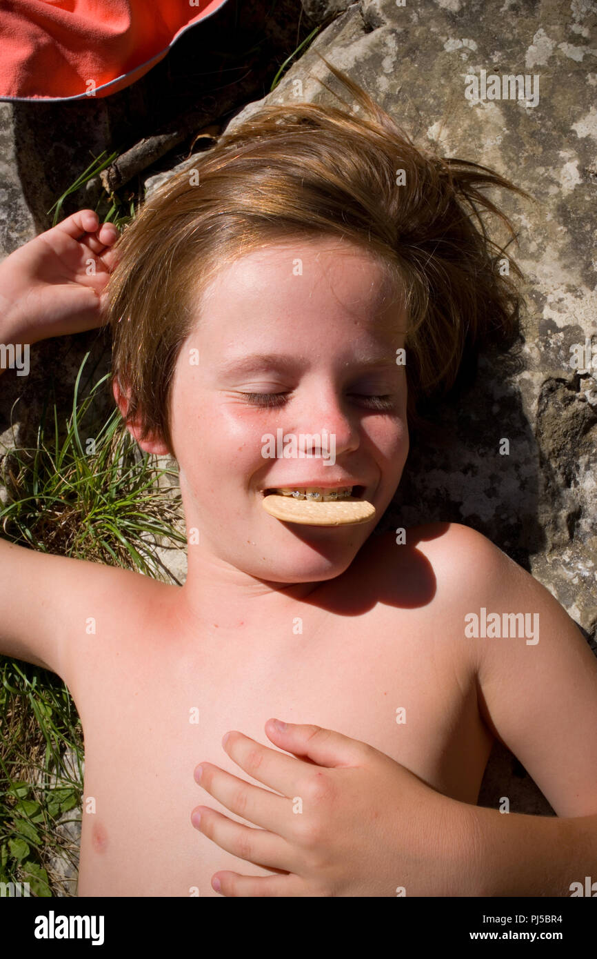 Nahaufnahme der elf Jahre alten Jungen Liegen in der Sonne mit seinen Augen geschlossen und lächelnd mit einem Keks zwischen seine Zähne mit Zahnspangen gehalten Stockfoto