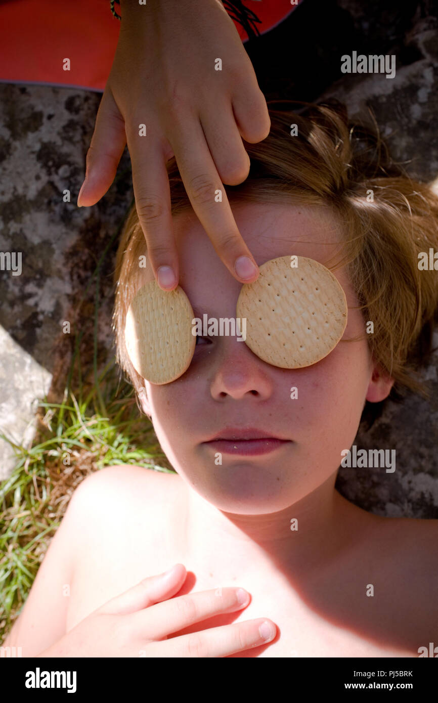 In der Nähe der elf Jahre alten Jungen Kopf und Schultern liegend im Schatten mit einem weiblichen Hand zwei runde Kekse über seine Augen Stockfoto