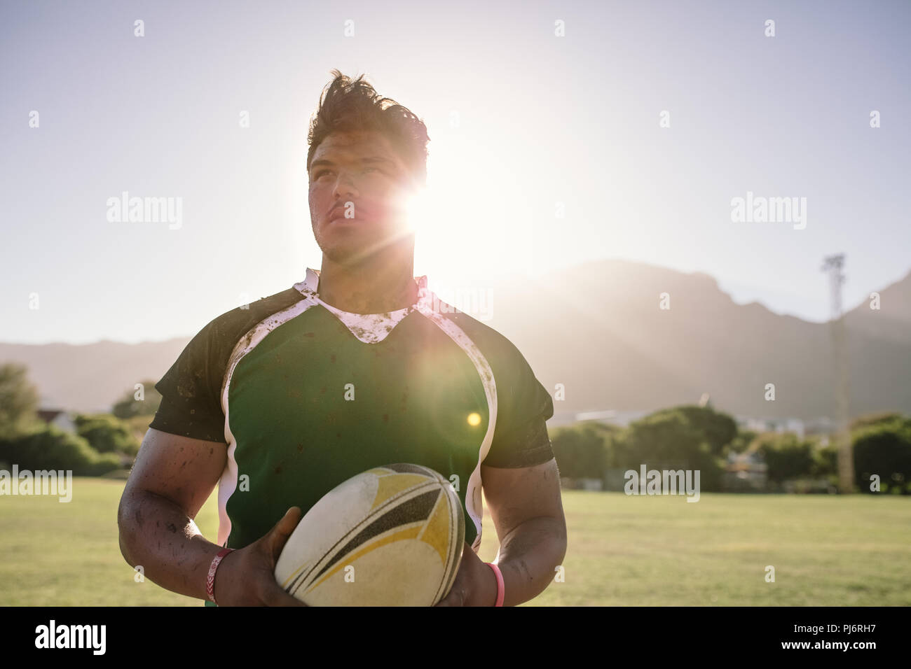 Junger Sportler, der einen Rugbyball mit schmutziger Uniform hält. rugbyspieler auf dem Platz mit hellem Sonnenlicht von hinten. Stockfoto