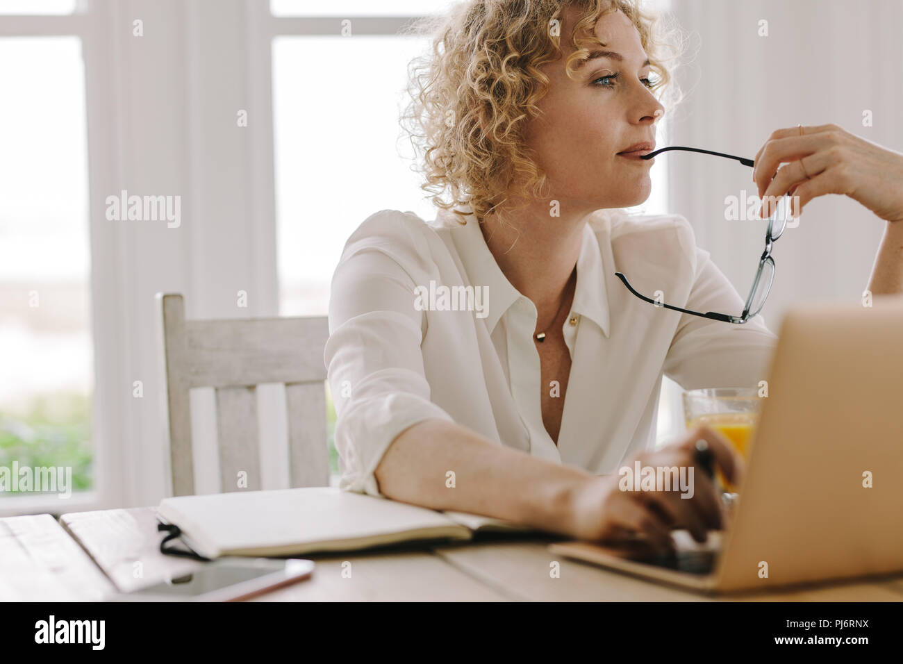 Frau tief zu denken, während auf dem Laptop zu Hause arbeiten. Frau sitzt zu Hause arbeiten mit Laptop und Tagebuch auf dem Tisch. Stockfoto