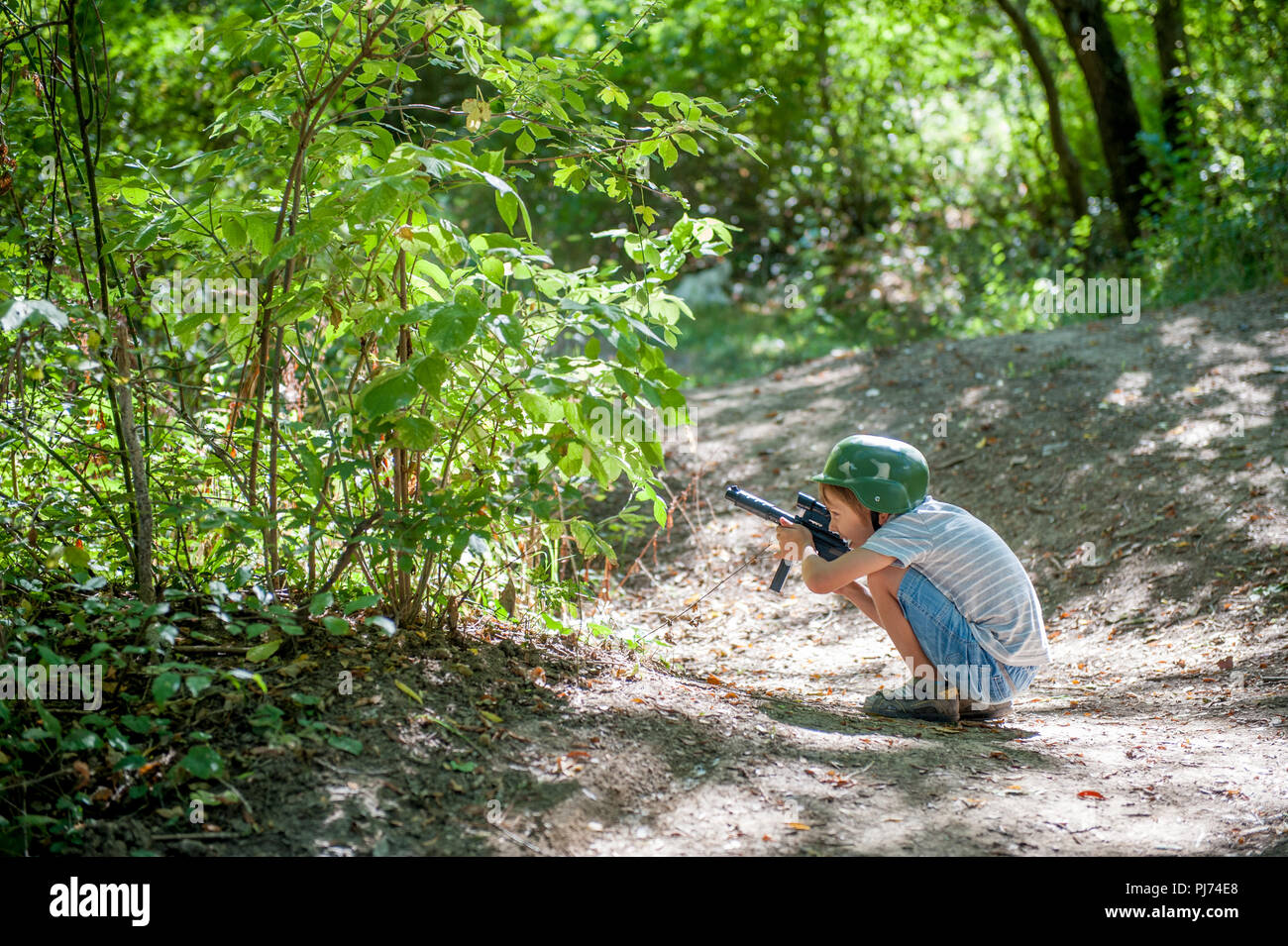 Lustige kleine kaukasischen Jungen in militärischen Helm versteckt hinter Bäumen mit spielzeugpistole in der Hand Stockfoto