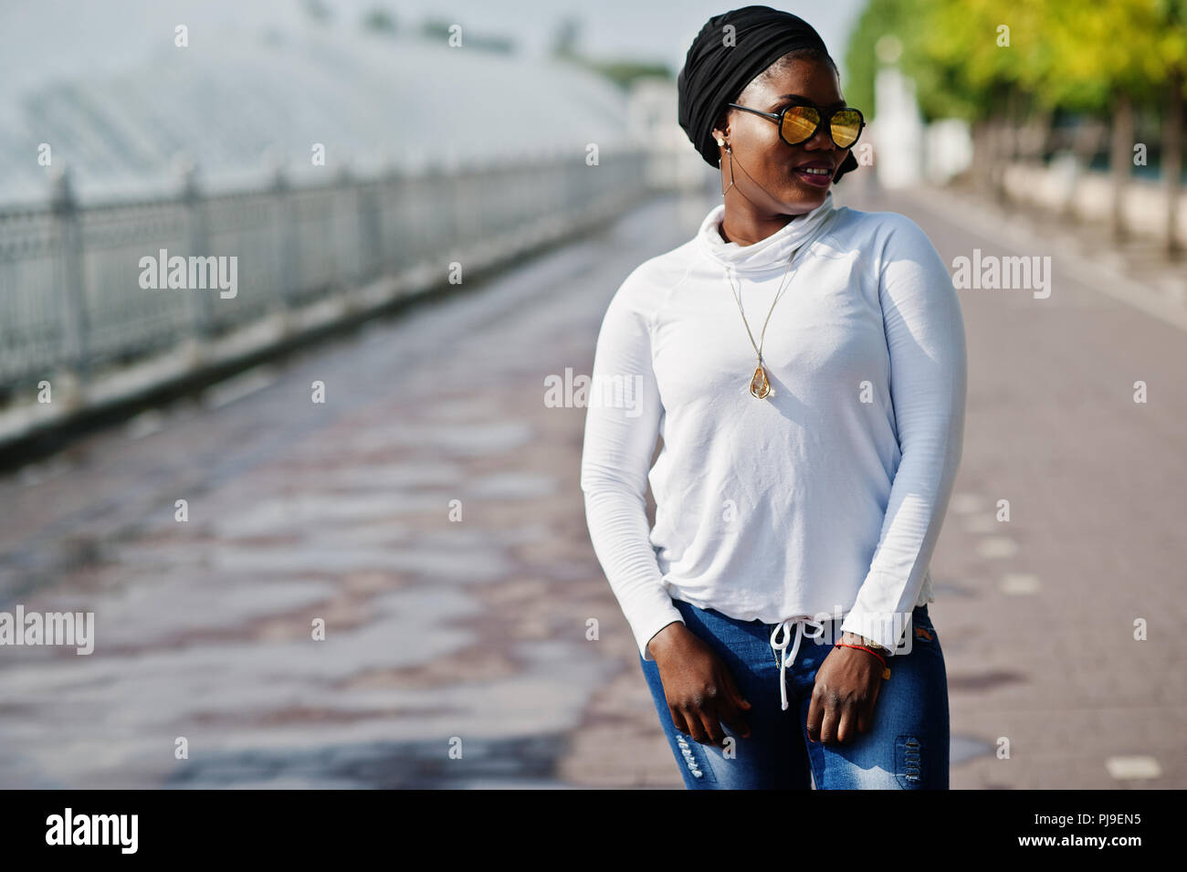 African Muslim Mädchen in der Sonnenbrille, schwarz Hijab, weißen Pulli und Jeans stellte im Freien gegen Brunnen. Stockfoto